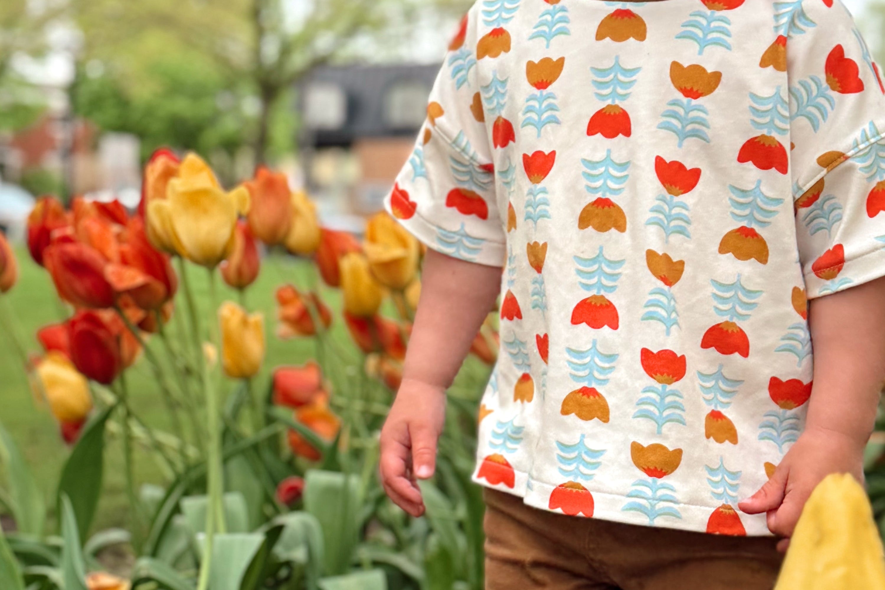 Child wearing a colorful shirt with floral patterns standing in a garden with tulips.